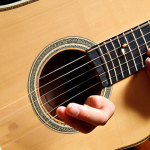 **Image Prompt 1:**
    "A close-up shot of a young Vietnamese beginner's hands on an acoustic guitar with nylon strings. Their fingertips appear slightly red, indicating initial finger pain from practice. On a nearby wooden surface, essential beginner guitar accessories are neatly arranged: a smartphone displaying a guitar tuner app, a capo, a few guitar picks of various thicknesses, and a small metronome. The setting is a warm, inviting practice space, bathed in soft light, conveying a sense of diligent effort and the foundational steps of learning guitar."